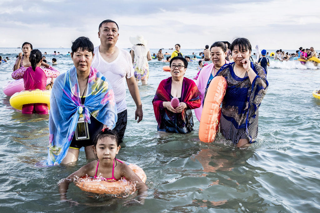 Nombre de Chinois ne sont encore jamais allés à la plage et beaucoup ne savent pas nager. Alors, quand ils vont à la mer, c’est pour la contempler, apprivoiser son rythme en trempant leurs pieds dans l’eau, se recouvrir de sable, faire des photographies au smartphone…
 Epiphénomène du développement du tourisme intérieur, les pratiques de la plage en Chine dessinent ainsi l’émergence d’une nouvelle activité de loisirs où les attitudes, tenues vestimentaires et accessoires de bain parlent d’eux-mêmes avec humour. 
Irrésistibles anti-héros du bord de mer, ils s’immergent ou déambulent arborant face-kini et vêtements anti-uv pour se protéger du soleil et surtout ne pas paraître bronzés, bouées multicolores autour de la taille ou du cou, pantalons retroussés pour aller dans l’eau jusqu’aux genoux, ombrelles… autant de pratiques insolites et joyeuses pour nos regards occidentaux !
Many Chinese have never been to the beach before and many do not know how to swim.
So when they go to the sea, it's to contemplate it, to tame its rhythm by dipping their feet in the water, to cover themselves with sand, to take photographs with a smartphone.
Epiphenomenon of the development of domestic tourism, beach practices in China are thus shaping the emergence of a new leisure activity where attitudes, clothing and bathing accessories speak for themselves with humor. 
Like anti-heroes by the seaside, they immerse themselves or walk around while wearing face-kini and anti-UV clothes to protect against the sun and especially not to look tanned, multicolored swim rings around the waist or neck, rolled up trousers to go in the water up to the knees, umbrellas... so many unusual and joyful practices for our Western eyes!